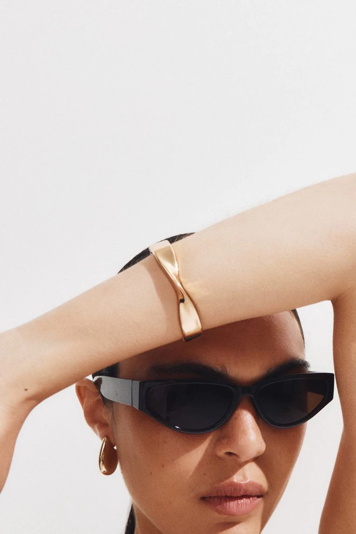Close-up portrait of a model wearing black sunglasses and a gold cuff bracelet against a plain white background