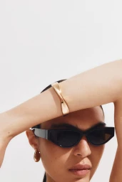 Close-up portrait of a model wearing black sunglasses and a gold cuff bracelet against a plain white background