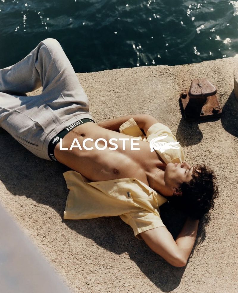 Man reclining shirtless by the water in an open yellow shirt, grey trousers, and Lacoste underwear waistband.