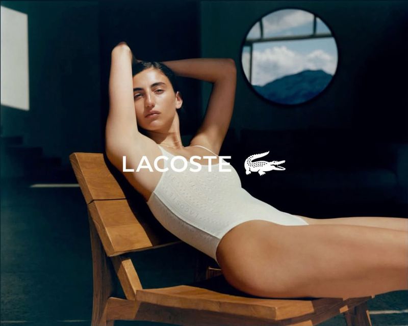 Model Paola Manes reclining on a wooden chair wearing a white one-piece bodysuit in front of a circular window.