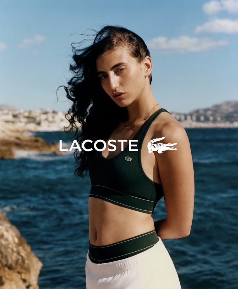 Model wearing a dark green Lacoste sports bra and white bottoms posing against a rocky coastal sea backdrop.