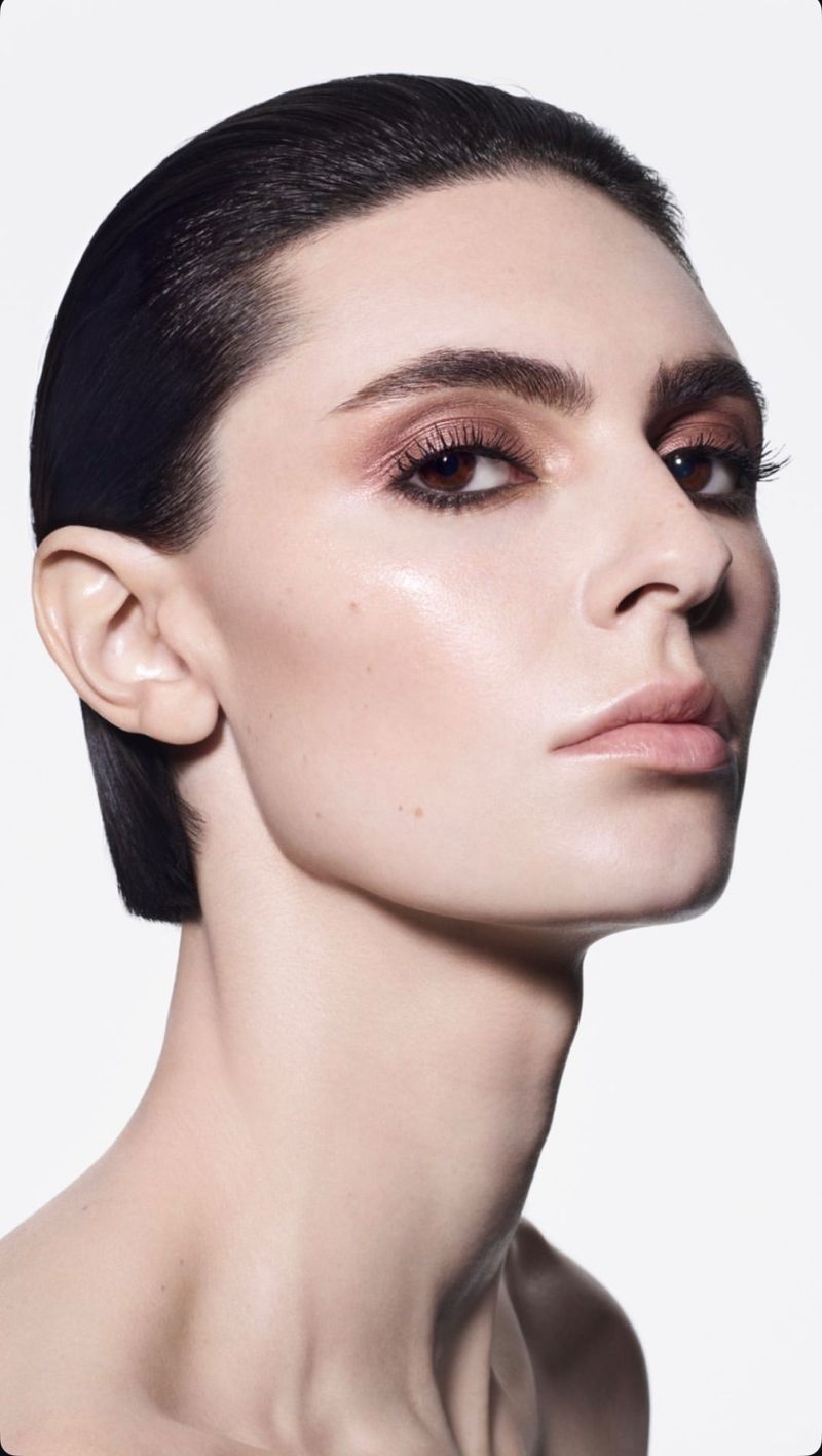 Close-up beauty portrait of a model with slicked-back dark hair, dewy skin, and smoky bronze eyeshadow against a white background.