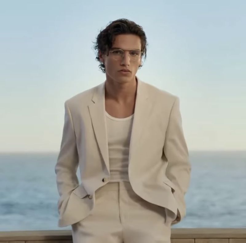 Model Morgan Sahadow wearing wire-rim glasses and an open cream tailored suit overlooking an ocean horizon.