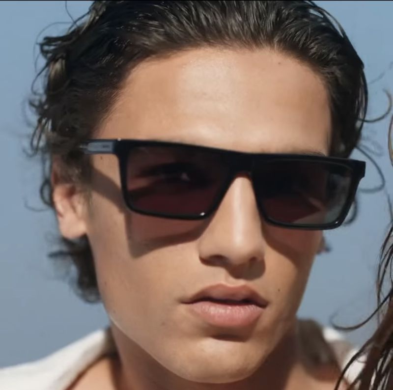 Close-up portrait of a man wearing black rectangular sunglasses with dark lenses outdoors under a clear blue sky.