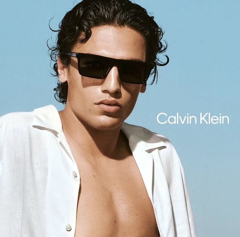 Model Morgan Sahadow wearing dark Calvin Klein rectangular sunglasses and an unbuttoned white shirt against a clear blue sky.