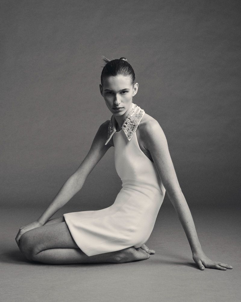 Model kneeling barefoot in a white sleeveless mini dress with an embellished collar against a neutral studio backdrop.