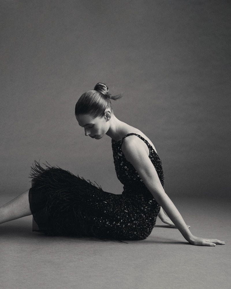 A woman in profile with a sleek bun sits on the floor wearing a black sequined cocktail dress with feather trim on a seamless backdrop.