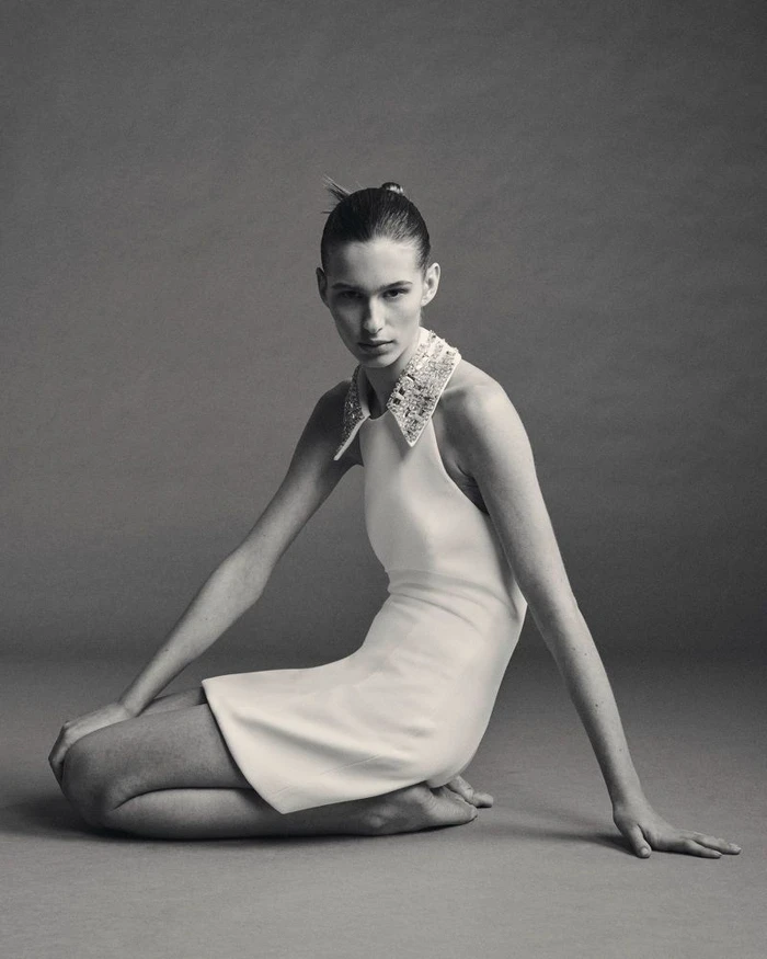 Model kneeling barefoot in a white sleeveless mini dress with an embellished collar against a neutral studio backdrop.