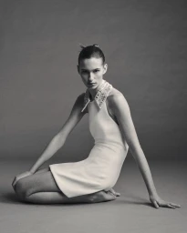 Model kneeling barefoot in a white sleeveless mini dress with an embellished collar against a neutral studio backdrop.