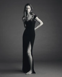 Model posing barefoot in a floor-length black evening gown with embellished neckline against a grey studio backdrop.