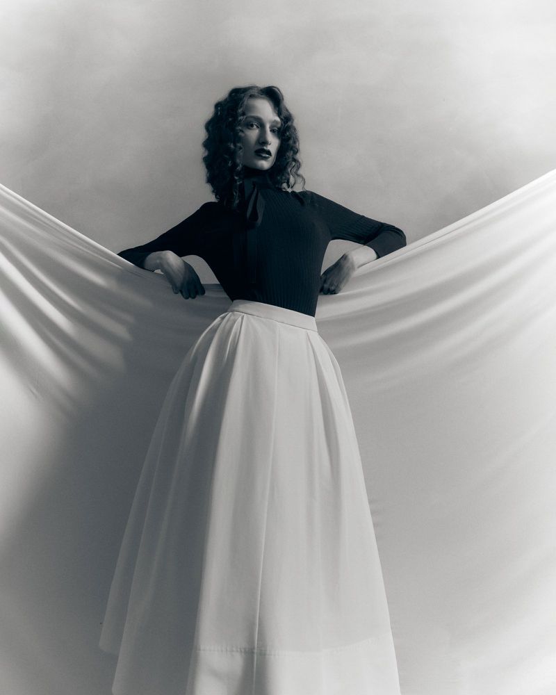 Woman with dark curly hair in a black long-sleeve top and flowing white maxi skirt holding draped white fabric behind her in a studio.