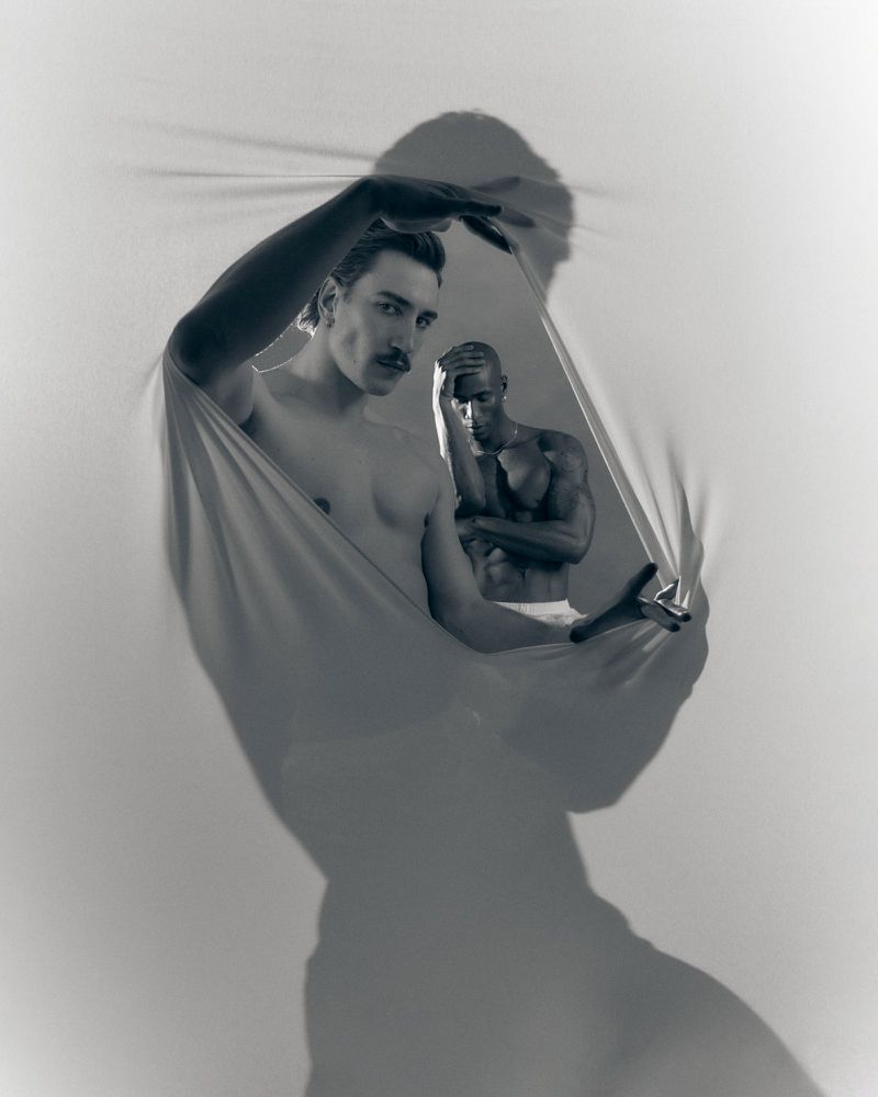 Shirtless man with mustache stretching an opening in a white fabric screen with another shirtless man behind him in monochrome.