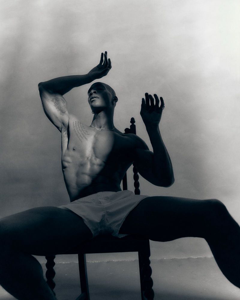 Monochrome portrait of a shirtless man seated on a carved wooden chair, striking a theatrical pose with raised arms against a cloudy sky.