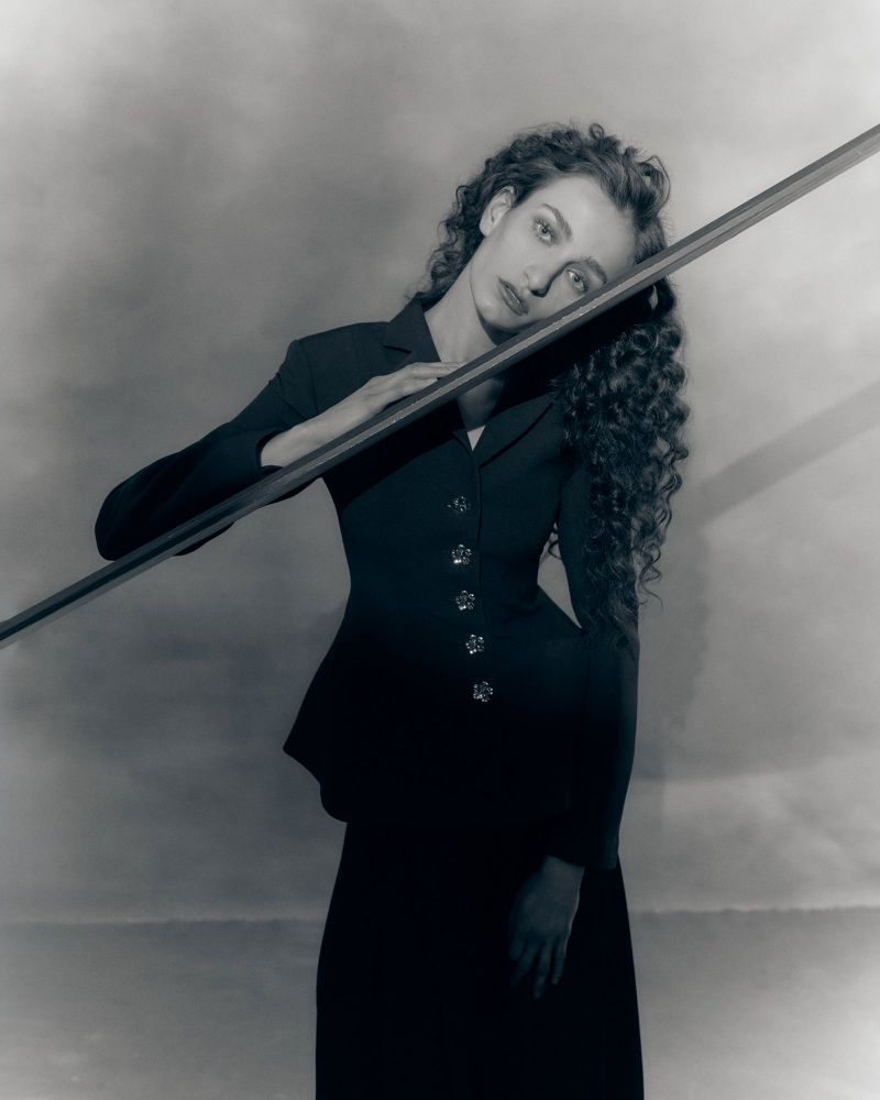 Model with long curly hair wearing a tailored black blazer leaning against a diagonal beam in monochrome portrait.