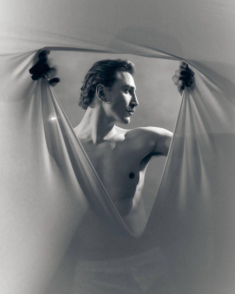 Shirtless man looking to the side while parting draped sheer fabric with both hands in a monochrome studio portrait.