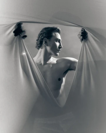 Shirtless man looking to the side while parting draped sheer fabric with both hands in a monochrome studio portrait.