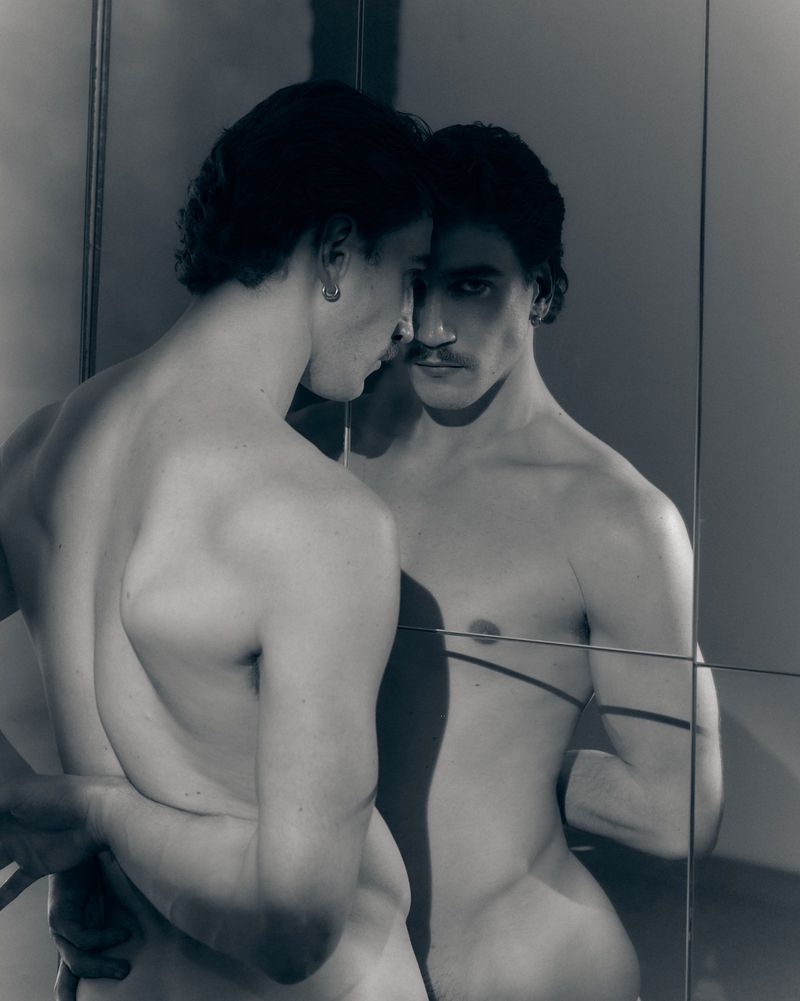 Monochrome portrait of a shirtless man with a mustache gazing into a grid of mirror panels, revealing his reflection.