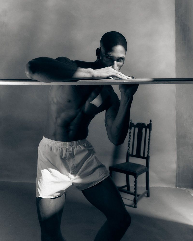 Black man posing shirtless in white boxer shorts behind a horizontal wooden plank in a studio setting.
