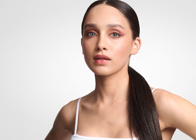 Close-up beauty portrait of a woman with slicked-back dark ponytail and soft neutral makeup against a plain white studio background.