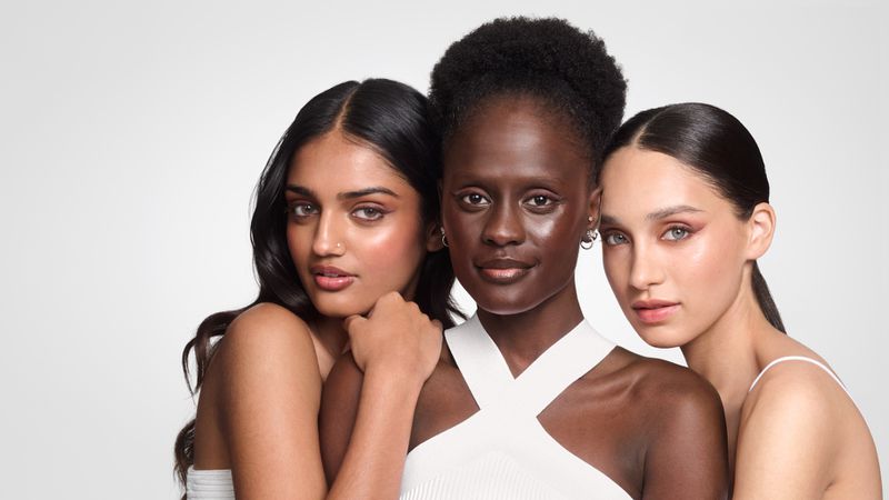 Three women of diverse skin tones posing closely together in white tops against a bright studio background.