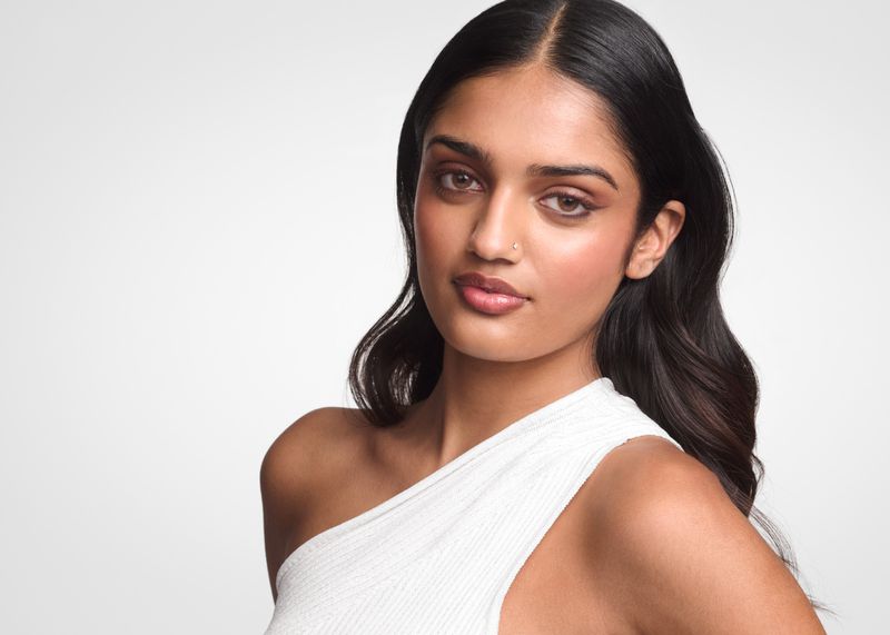 South Asian woman with dark wavy hair and radiant makeup wearing a white one-shoulder top against a neutral studio backdrop.
