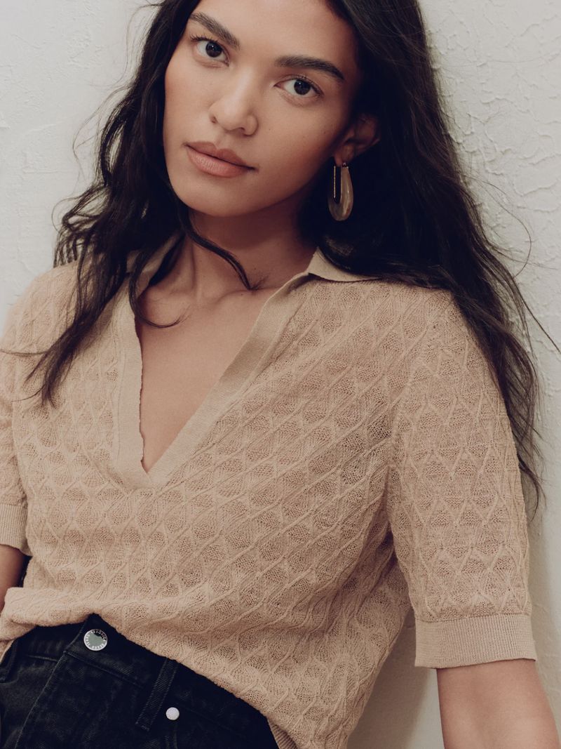 Woman posing in a textured beige polo shirt with short sleeves, wearing hoop earrings, against an off-white wall in a medium shot.