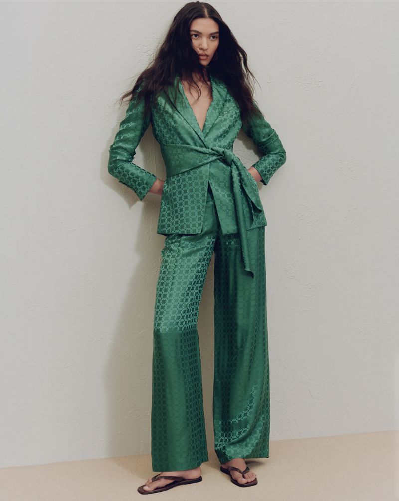 Elegant woman in a patterned emerald green blazer and wide-leg pants, posing in front of a neutral wall in a fashion shot.