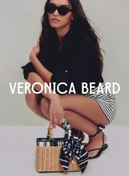 Model crouching wearing black shirt and sunglasses with striped shorts, holding a woven basket bag for Veronica Beard.