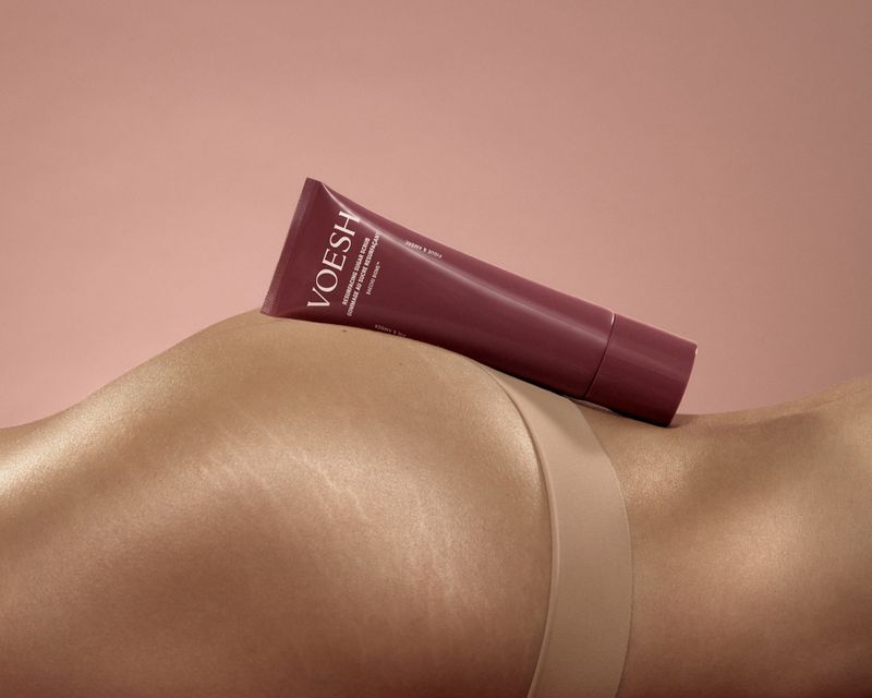 A burgundy tube of VOESH resurfacing sugar scrub resting along the curve of a model's hip against a warm pink background.