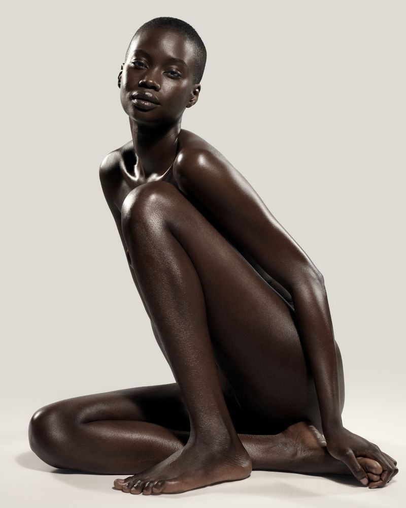 Model Fatou Jobe seated in a crouched pose with luminous glowing skin against an off-white seamless background.
