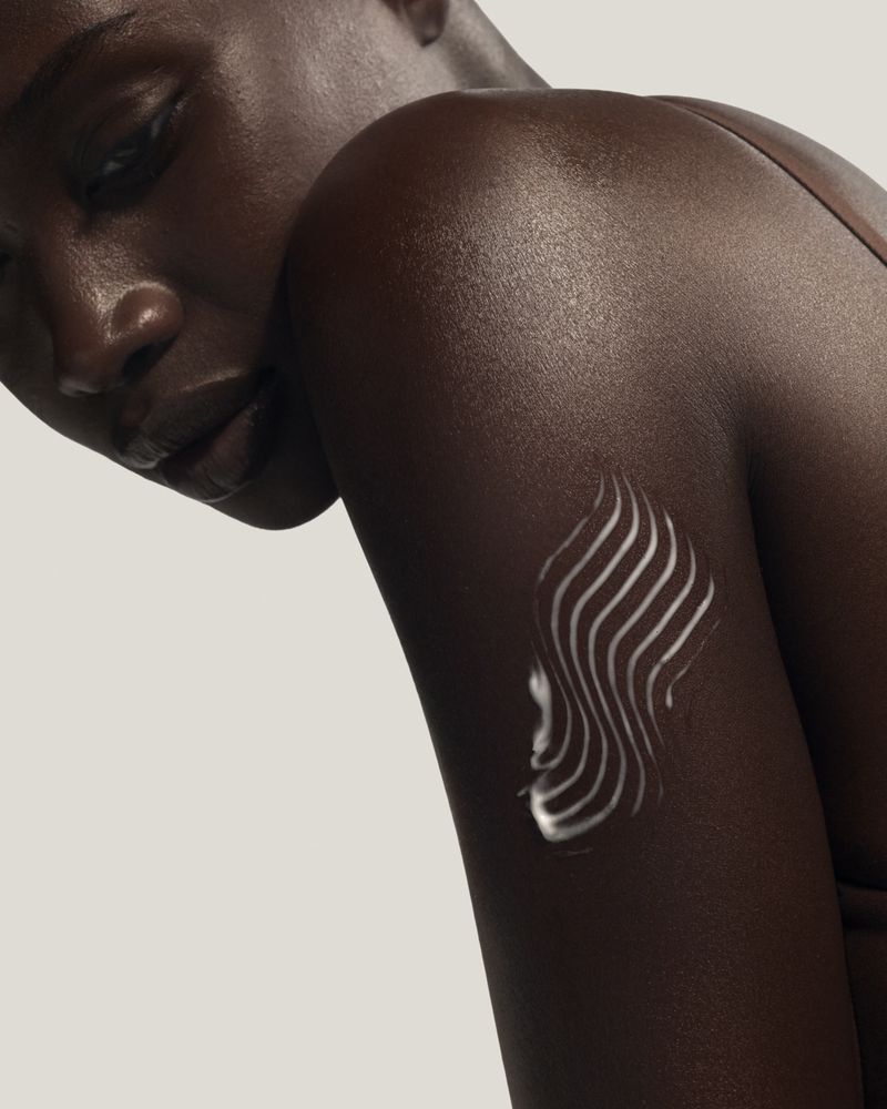 Close-up of a Black woman's shoulder and arm featuring a textured wavy swatch of white body cream against a neutral backdrop.