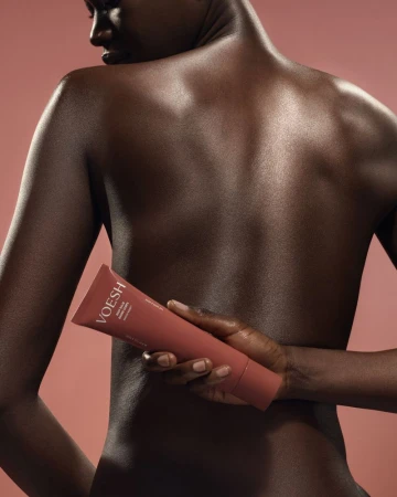 Black model holding a rose-colored VOESH lotion tube behind her bare back against a solid pink studio background.