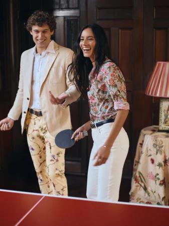 A smiling man and woman playing table tennis indoors, wearing floral and tailored resort wear beside a red ping pong table.
