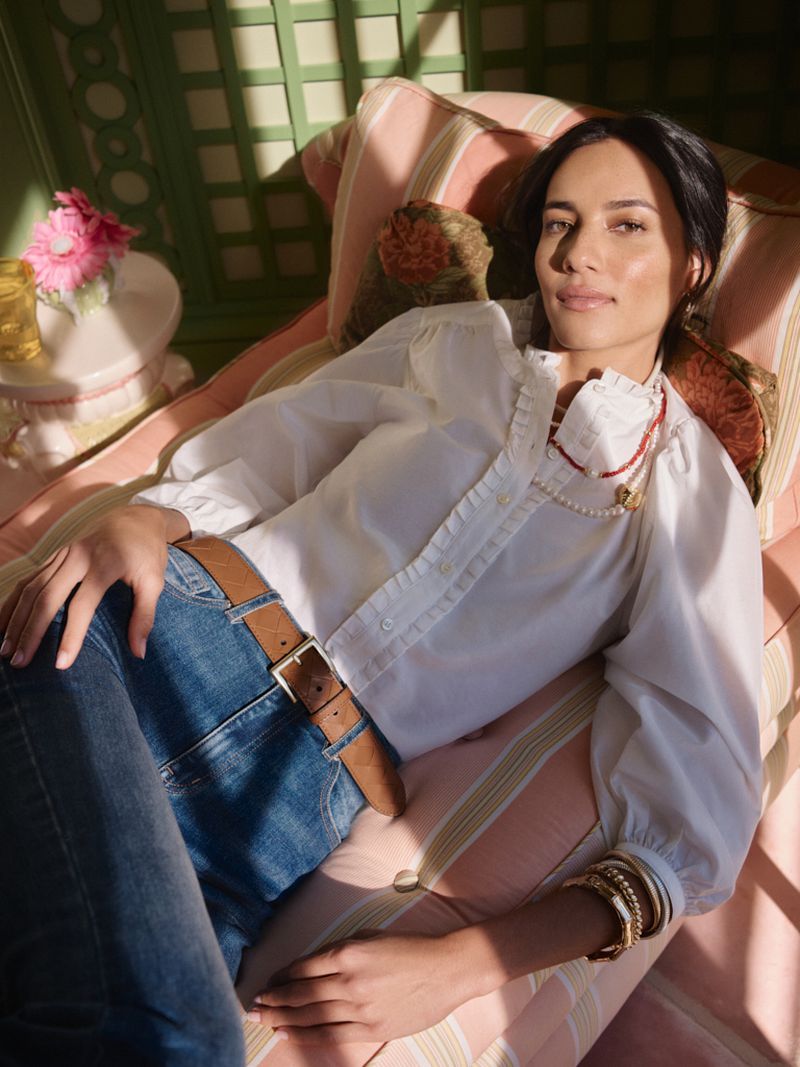 Woman reclining on a pink striped chaise lounge wearing a white ruffled blouse, blue denim jeans, and a brown leather belt in sunlight.