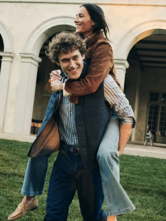 A smiling man gives a piggyback ride to a smiling woman on a lawn in front of an estate with arched colonnades.