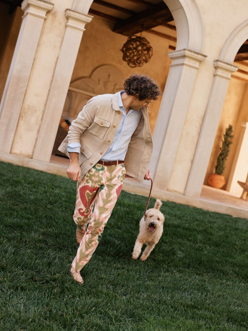 A man with curly hair walks a dog on a leash across a green lawn, with architecture in the background featuring archways and planters.