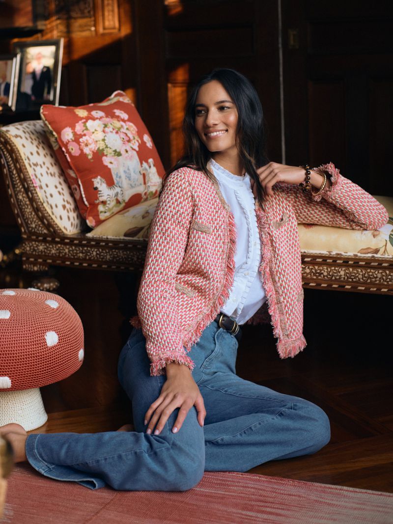 Woman seated on the floor wearing a pink fringe tweed jacket, white ruffled blouse, and blue jeans beside a patterned settee.