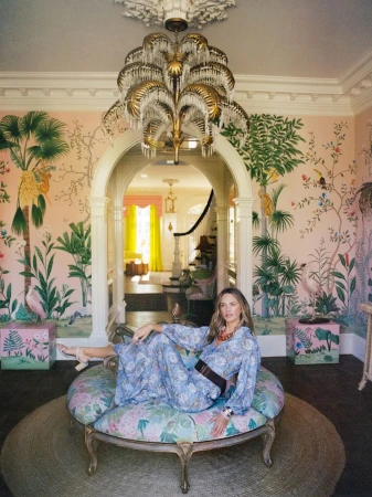 Woman in a belted floral blue dress reclining on a round upholstered ottoman in a room with pink tropical wallpaper.