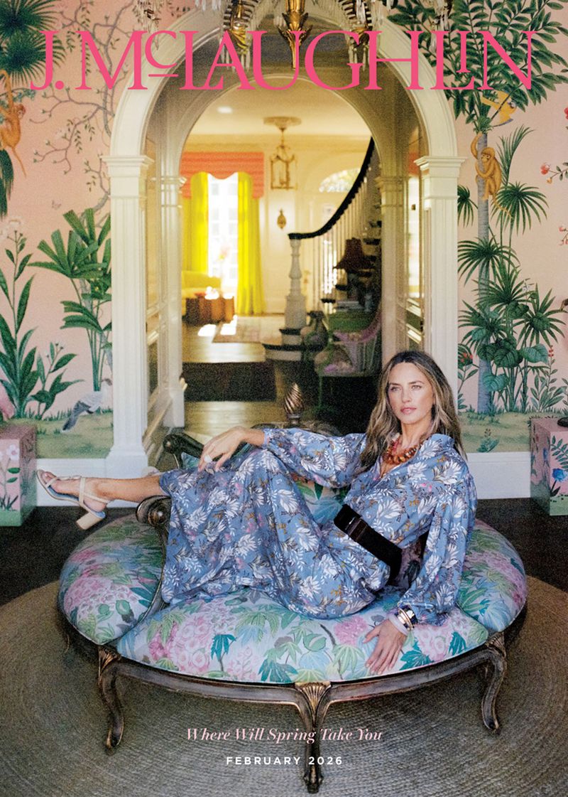 A woman in a blue floral print maxi dress reclines on an ornate round upholstered settee in an entryway with tropical wallpaper.