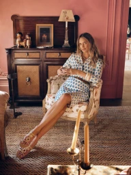 A woman in a blue and white patterned shirt dress sits in a vintage armchair against pink walls in a warm interior setting.