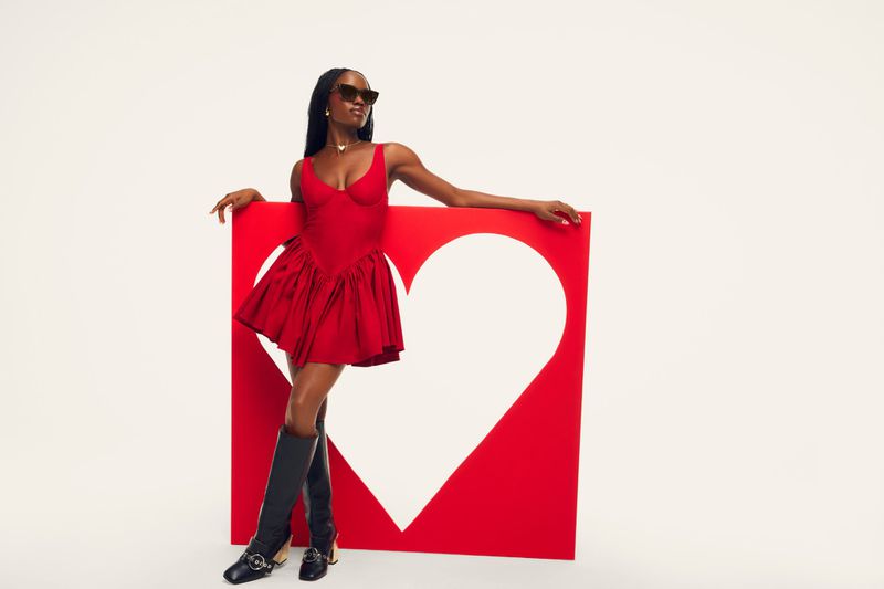 A woman in a red minidress, dark sunglasses, and black knee-high boots posing beside a large red heart cutout prop on a white background.