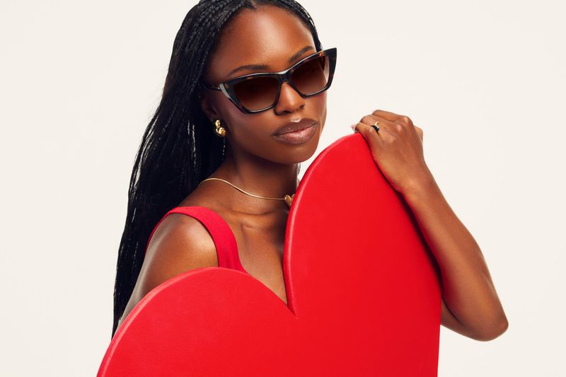 A Black woman with long braided hair wearing tortoiseshell cat-eye sunglasses holds a large red heart prop against a plain light backdrop.