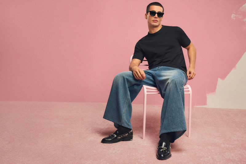 Man wearing black sunglasses, a black t-shirt, and wide-leg blue jeans seated on a pink chair against a pink background.