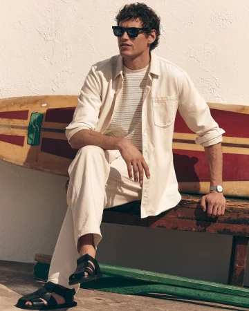 Model Paul Hupfer sitting on a wooden bench with a vintage surfboard wearing an off-white shirt, cream pants, sunglasses, and sandals.