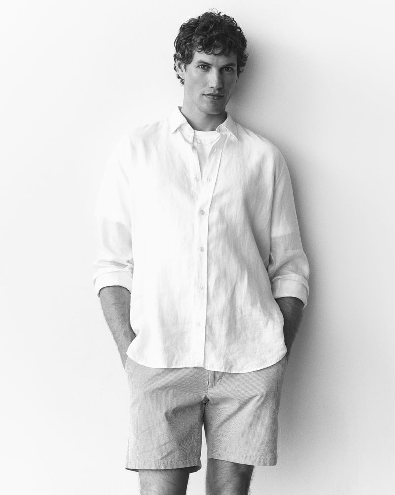 Model Paul Hupfer wearing a white linen button-down shirt and shorts standing against a white studio background.