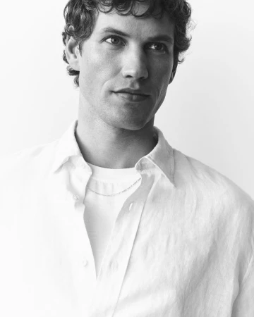 Black and white portrait of a man with curly hair wearing an open white shirt over a white tee against a white backdrop.