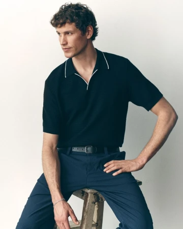 Model Paul Hupfer seated on a wooden ladder stool wearing a black collared polo and blue trousers against a white background.