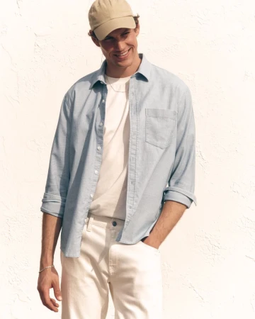 Model Paul Hupfer wearing a light blue button-down shirt, beige baseball cap, and white pants against a textured white wall.