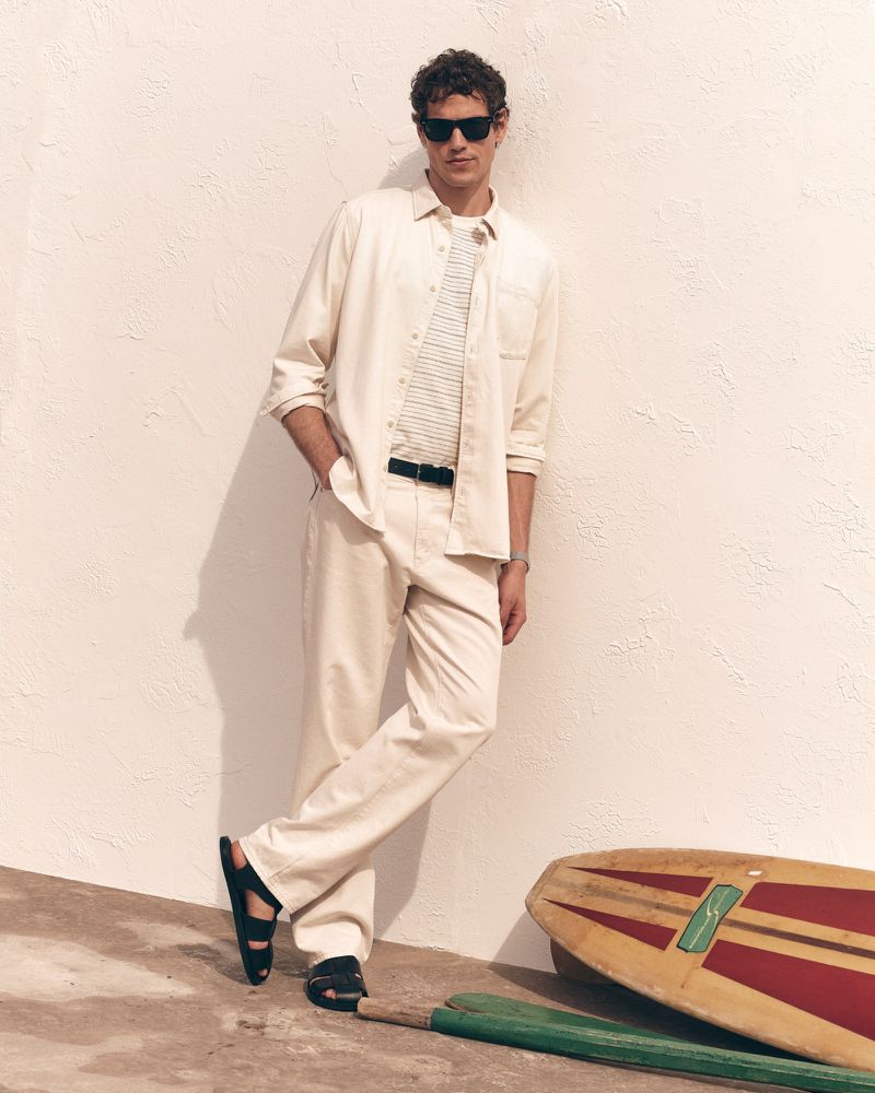 Model Paul Hupfer leaning against a wall wearing a cream shirt, trousers, and sunglasses beside a vintage surfboard.