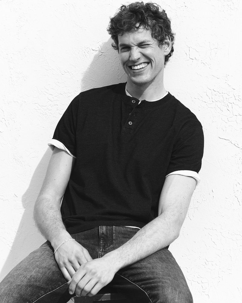 Model Paul Hupfer smiling and winking against a white textured wall in a black henley shirt and jeans for Mavi.